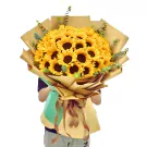 A bouquet of 36 sunflowers wrapped in brown and green paper, held by a person.