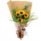 Bouquet of sunflowers in brown paper with green foliage and brown ribbon.