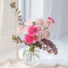 Pink roses, carnations, and gerbera daisies in a round glass vase with eucalyptus branches.