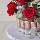 Red roses, carnations, and eucalyptus surround a cake decorated with rolled Vietnamese banknotes and a silver ribbon.