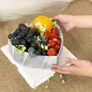 Heart-shaped fruit box with grapes, strawberries, and an orange, decorated with flowers and greenery.