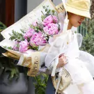 A bouquet of pink peonies and baby's breath wrapped in decorative paper held by a person in a yellow hat.