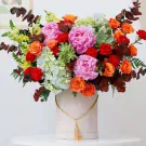 A bouquet of pink peonies, orange roses, red carnations, hydrangeas, and succulents in a beige hat box.