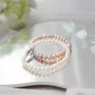Three delicate pearl bracelets in white, pink, and peach colors, reflecting on a mirrored surface.