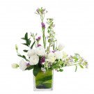 White roses, statice, and blue star flowers arranged in a square glass vase with green foliage.