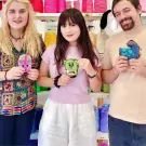 Three people hold up colorful punch needle creations in a craft workshop.