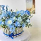 Blue and white floral arrangement in a box decorated with rolled banknotes, tied with a blue ribbon.