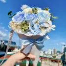 A cone-shaped bouquet of pastel blue and white roses, tied with a blue ribbon, held against a bright blue sky.
