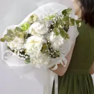 White peonies in a bouquet with eucalyptus and baby's breath, wrapped in white mesh, held by a person in a green dress.