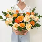 Basket of orange and peach roses with greenery, held by a person wearing a white floral top and jeans.