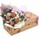 Bouquet of pink roses, yellow chrysanthemums, and dark pink pom-pom flowers in a cardboard box.