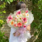 A person holds a bouquet of pink and orange tulips, David Austin roses, and gerbera daisies, wrapped in white paper.