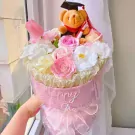 Graduation gift basket with a teddy bear wearing a cap and diploma, surrounded by pink and white flowers.