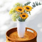 Bouquet of sunflowers and hydrangeas in a white vase on a rattan tray.