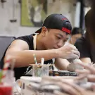 Man in a black cap and apron sculpts clay, working on a detailed piece.