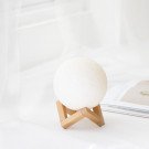 A white moon-shaped lamp with a textured surface sits on a wooden stand beside an open book.