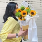 Woman in yellow shirt holding a bouquet of sunflowers, green flowers, and white daisies with a large white ribbon.