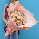 A bouquet of yellow and orange David Austin roses with grey-green foliage wrapped in peach and white paper, tied with a pink ribbon.