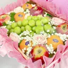 Fruit basket with green grapes, oranges, red apples, and pink roses, wrapped in pink mesh.