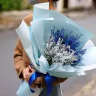 A person holds a bouquet of blue and silver dried flowers wrapped in light blue paper.