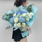 Bouquet of blue chrysanthemums, white roses, and greenery wrapped in light blue paper.