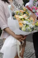 A person holds a delicate bouquet of peach roses, hydrangeas, and carnations wrapped in white paper with a newspaper insert.