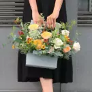 A person holding a light blue box of "Spring Morning" flowers with roses, sunflowers, and greenery.