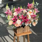Floral arrangement in a rustic wooden box, featuring pink lilies, roses, calla lilies, and greenery.