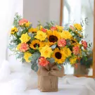 A bouquet of sunflowers, yellow roses, and orange-edged carnations in a burlap box.