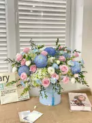 Light blue and pink floral arrangement in a cylindrical box, with books and cards on a burlap surface.