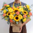 A bouquet of sunflowers, yellow roses, and orange carnations in a burlap box.