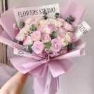 A bouquet of pink and white roses with purple accents, wrapped in lilac paper with a white ribbon.