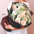 A hand holding a bouquet of cream roses, baby's breath, and daisies, wrapped in dark brown paper.