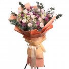 Bouquet of pink and purple roses, hydrangeas, and eucalyptus in orange and peach paper wrapping with a cream ribbon.