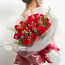 A bouquet of red roses with greenery and a red ribbon, held by a person in a pink dress.