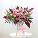 A large, elaborate floral arrangement with pink and cream roses, chrysanthemums, and gerbera daisies on a wooden stand.