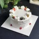 A white fruit cake topped with strawberries, grapes, and whipped cream, on a white cake board.