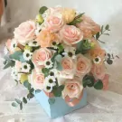 A light blue box filled with pink roses, peach carnations, and white daisies, accented with eucalyptus leaves.