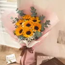 A bouquet of sunflowers and eucalyptus, wrapped in pink paper, tied with a brown ribbon.