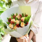 Bouquet of peach roses with red berries and green leaves, wrapped in white and green paper, tied with a peach ribbon.
