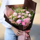A bouquet of pink roses and carnations wrapped in brown and pink paper, held by a person in a white top and blue jeans.