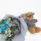 Teddy bear with sunglasses next to a bouquet of blue and white flowers.