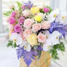 A floral arrangement with pink and yellow roses, white orchids, and purple accents in a wooden box.