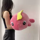 A person holds a large, pink, plush dinosaur toy with big eyes and a yellow tail.