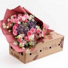 A bouquet of pink roses, purple hydrangeas, and other flowers in pink wrapping, displayed on a brown box.