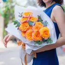 Woman holding a bouquet of vibrant orange and pink roses wrapped in white paper and tied with an orange ribbon.