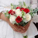 Bridal bouquet of red and white roses with greenery held by a person in a white dress.