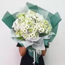 A bouquet of white hydrangeas and daisies, wrapped in green and white paper, held by a person.