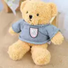 A light brown teddy bear wearing a blue knitted sweater with a patch on the chest sits on a tan surface.