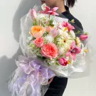 Beautiful bouquet of pink and orange roses, tulips, and white flowers wrapped in sheer paper with a purple ribbon.